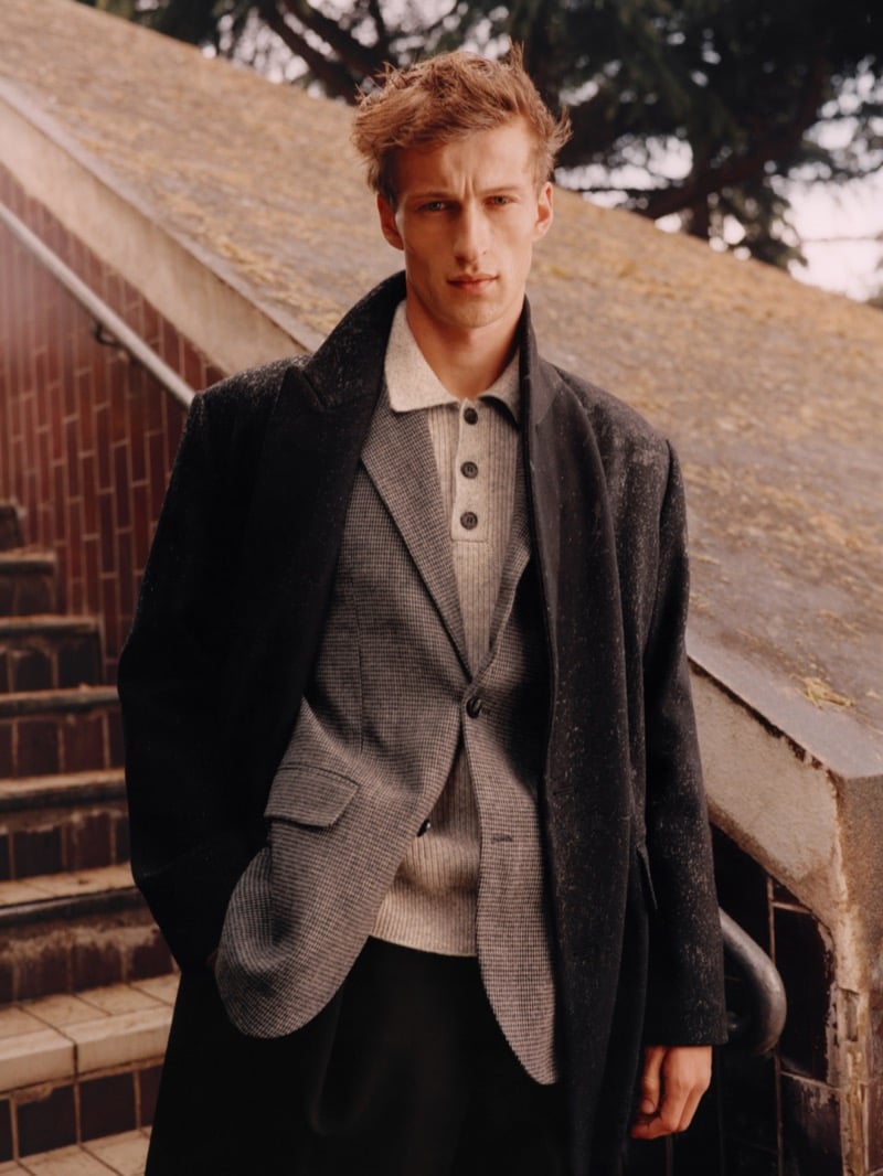 Vasko Luyckx on a tiled outdoor staircase in a dark textured overcoat over a gray checked blazer and a ribbed knit polo