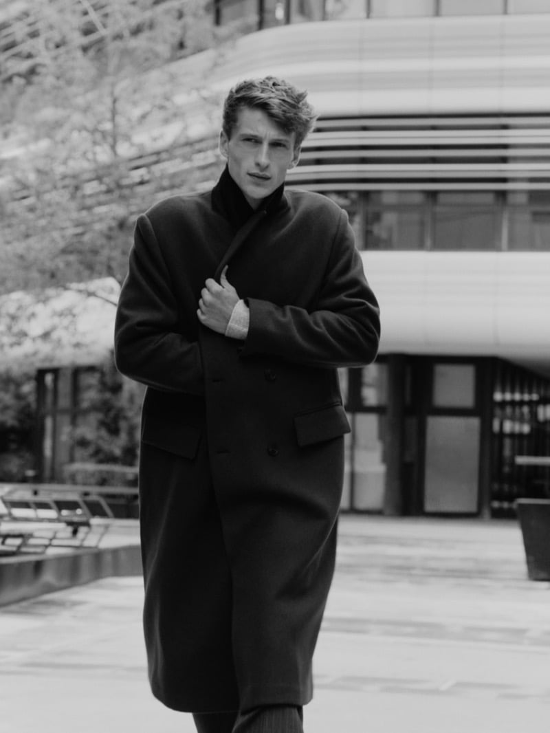 Vasko Luyckx in a black-and-white photo walking past a modern building in a long double-breasted overcoat, one hand drawing the lapel across his chest