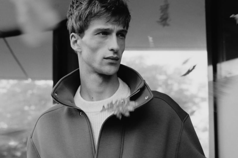 Vasko Luyckx in a black-and-white photo wearing a zip-front high-collar blouson over a waffle-knit sweater, leaves drifting past in the foreground