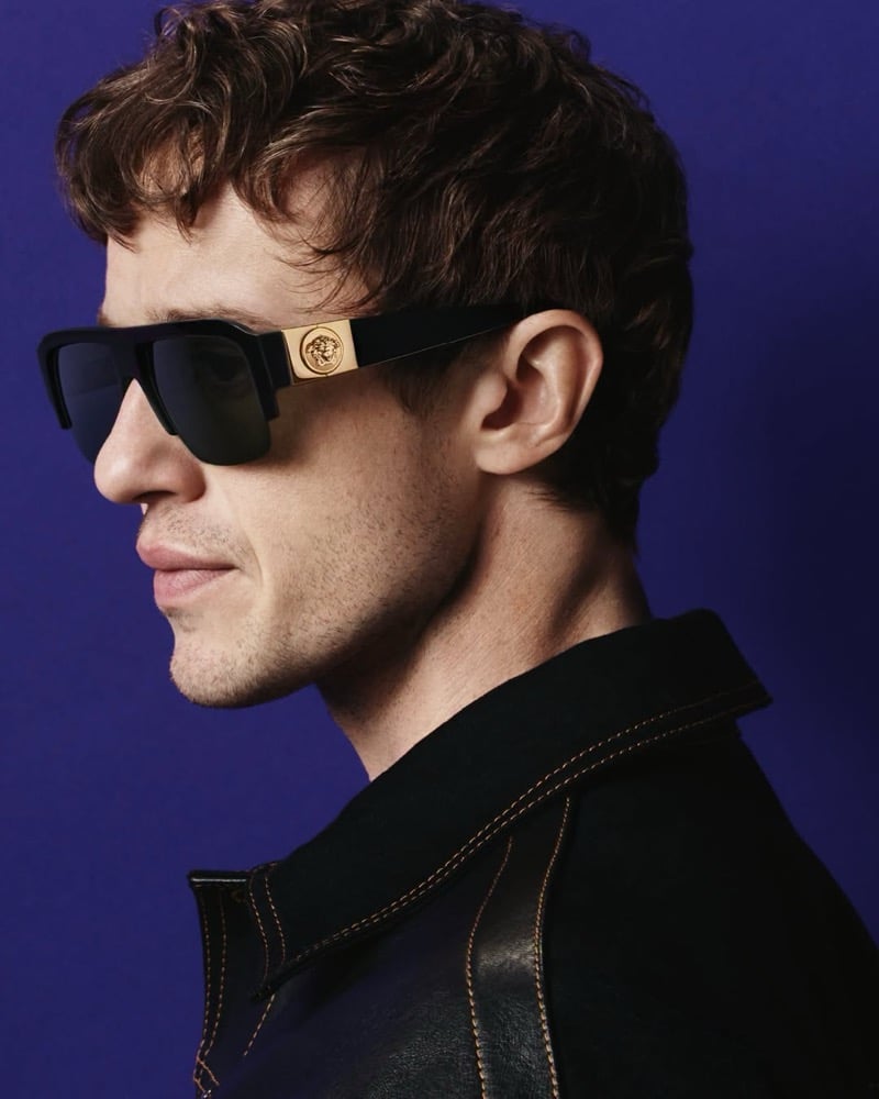 Joseph Quinn in profile wears black half-rim sunglasses with a gold Medusa temple plaque and a dark topstitched jacket against a violet backdrop for Versace's fall 2026 eyewear campaign.