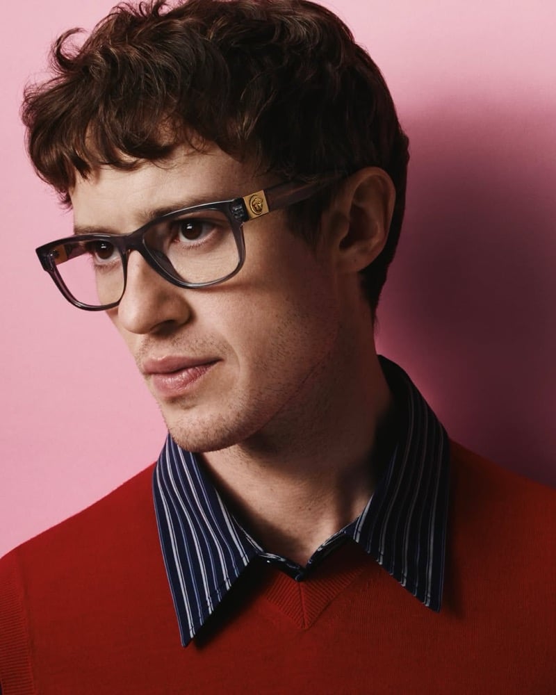 Joseph Quinn looks off to the side in smoky gray optical frames with a gold Medusa detail, a red V-neck sweater, and a striped collared shirt against a pink backdrop for Versace's fall 2026 eyewear campaign.