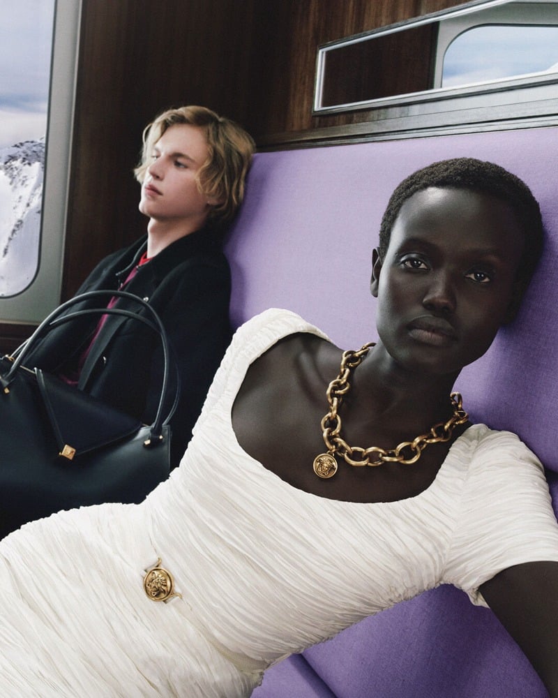 Yar Aguer wears a white dress and gold chain necklace beside Max Stenzel in a black coat holding a black handbag.