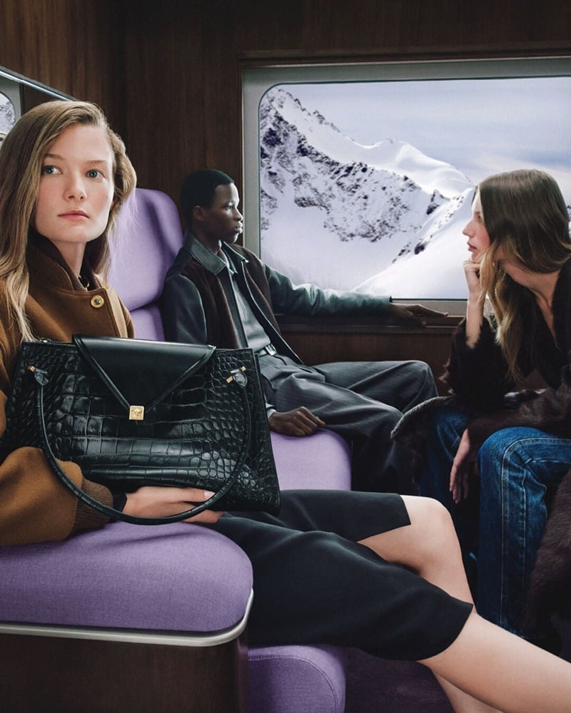 Reina Spoormaker holds a textured black handbag beside Micheal Okotie in a leather jacket and gray trousers, with Esme Cornelius seated opposite.