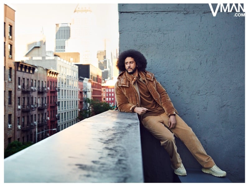 Colin Kaepernick sits on a rooftop ledge in a brown Moncler jacket and tan trousers.