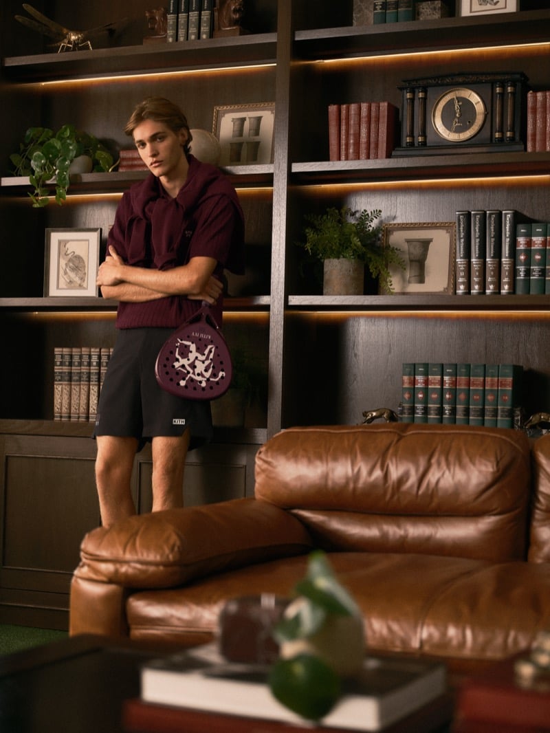 Warren Axelman standing with arms crossed in front of lit wood bookshelves in a burgundy polo with a sweatshirt knotted over his shoulders, black shorts, and a padel racket at his side