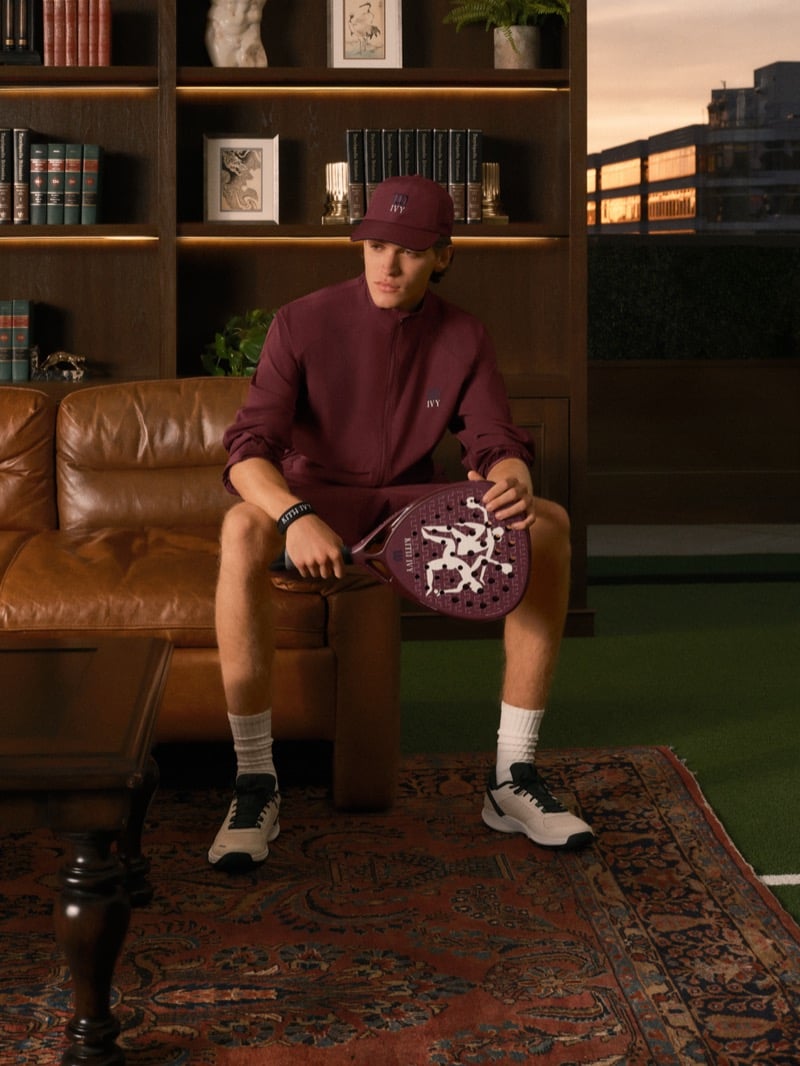 Warren Axelman seated on a leather sofa in a burgundy zip jacket, shorts, and cap, a padel racket with a graphic face across his knee, a sunset skyline in the window