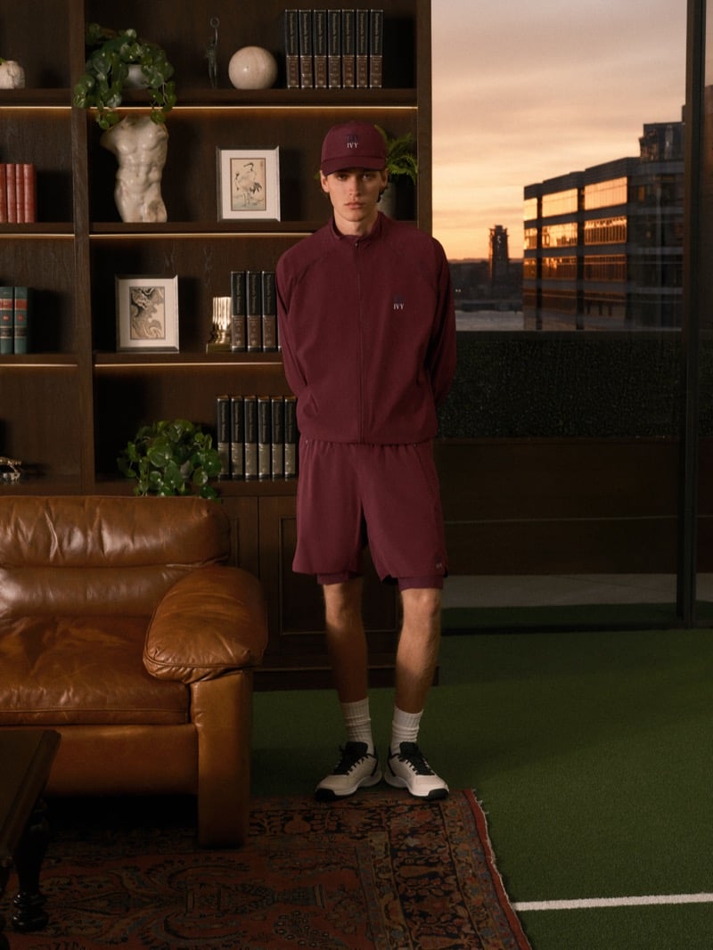 Warren Axelman standing with his hands behind his back in a burgundy zip jacket, matching shorts, a cap, and sneakers, with a sunset city view behind him