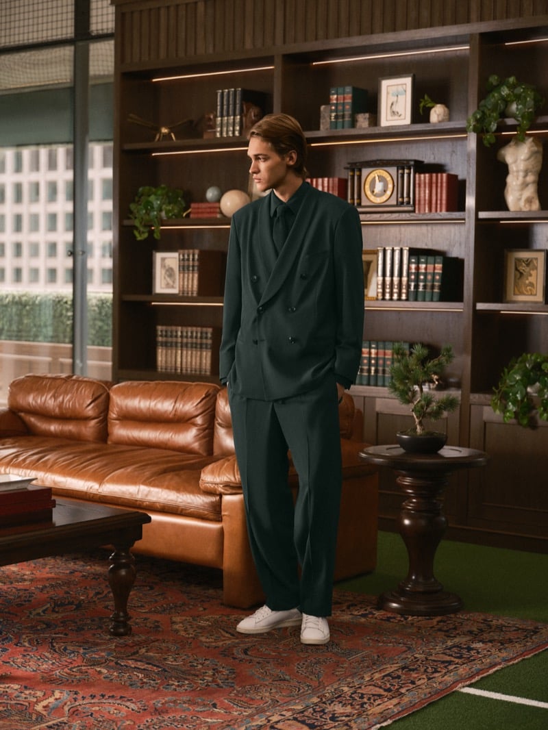 Warren Axelman standing in profile on a patterned rug in a green double-breasted suit with a tie and white sneakers, a leather sofa and bookshelves behind him