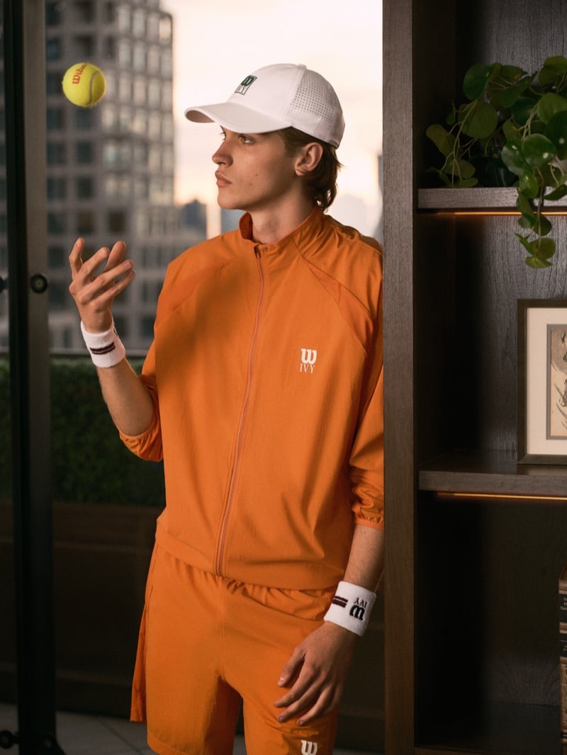 Warren Axelman tossing a tennis ball in a light cap, an orange zip jacket with a Wilson Ivy logo, shorts, and wristbands, a city window behind him