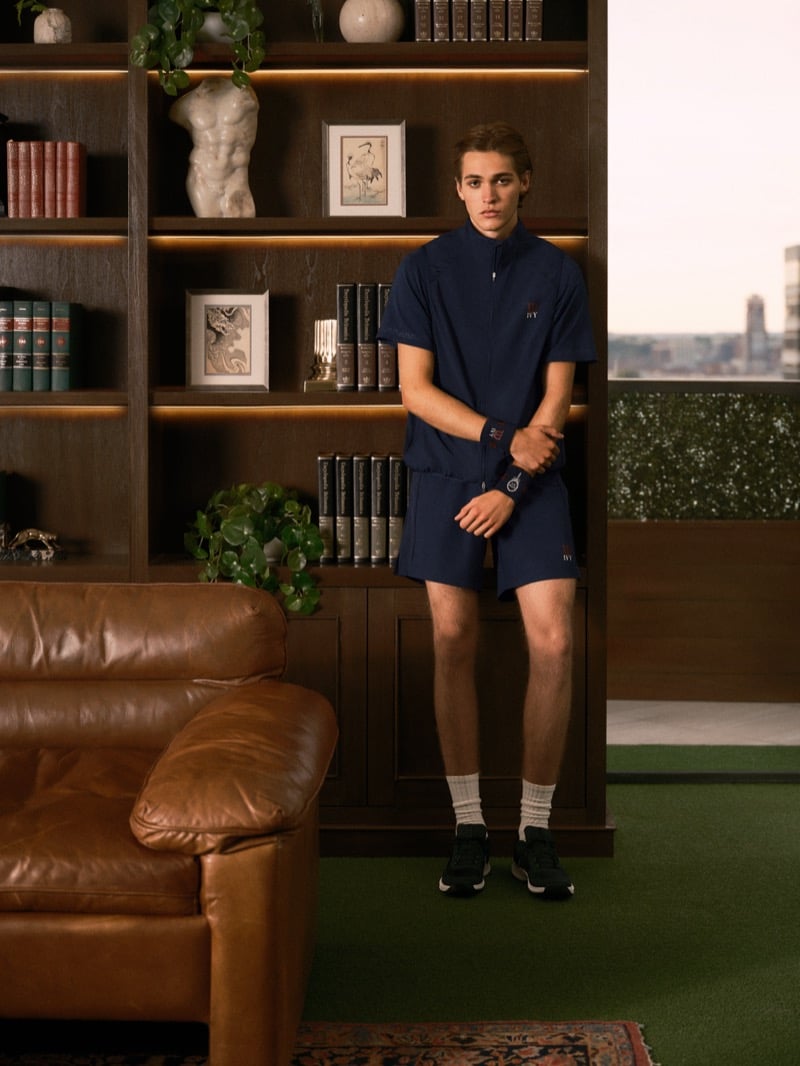 Warren Axelman standing beside a brown leather sofa in a short-sleeve zip top and matching shorts with wristbands, crew socks, and black sneakers, a city skyline in the window behind him