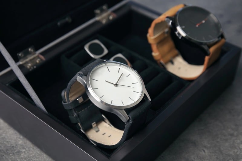 Two wristwatches on separate cushions in a black storage case with an open glass lid.
