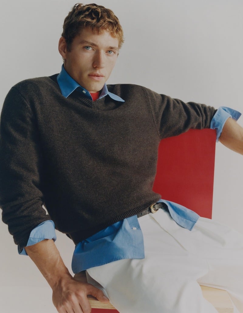 Valentin Humbroich sits in a red chair wearing a dark sweater over a blue shirt with white trousers.