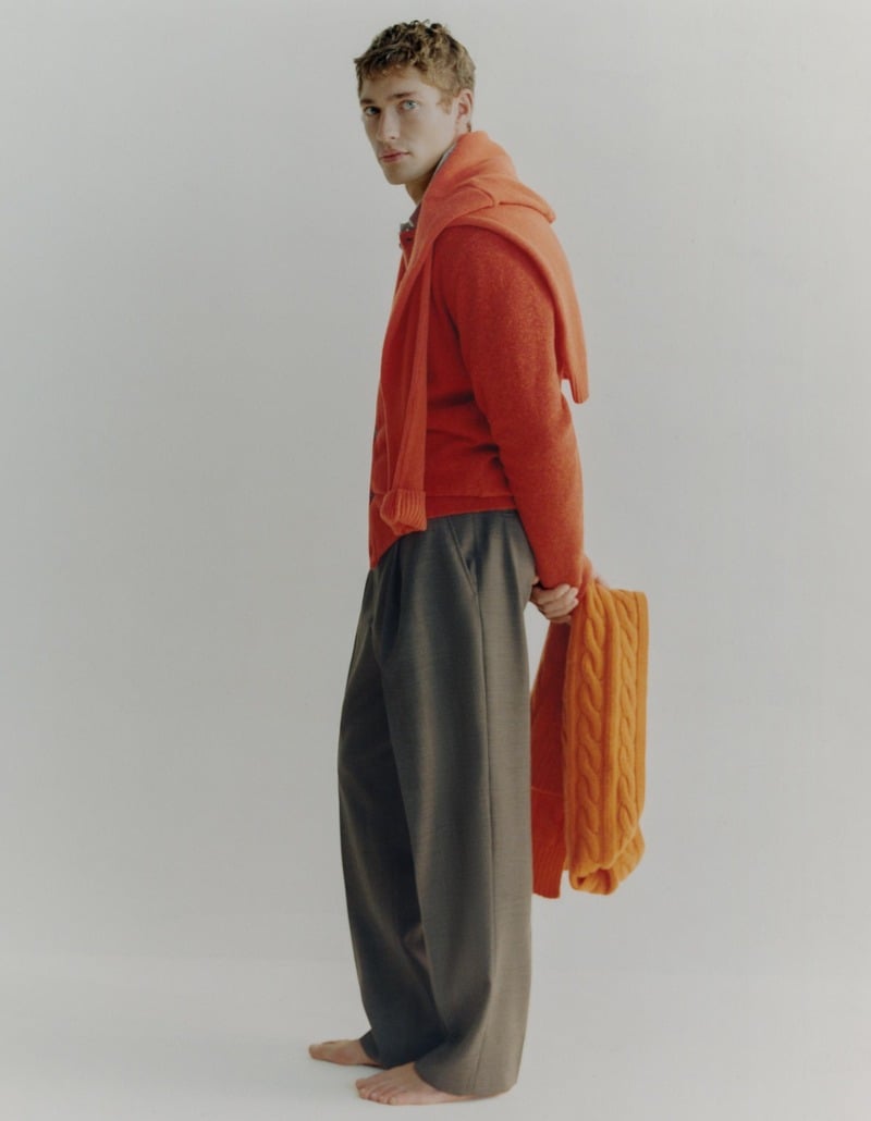 Valentin Humbroich stands barefoot in an orange cardigan and loose gray trousers, holding an orange knit.