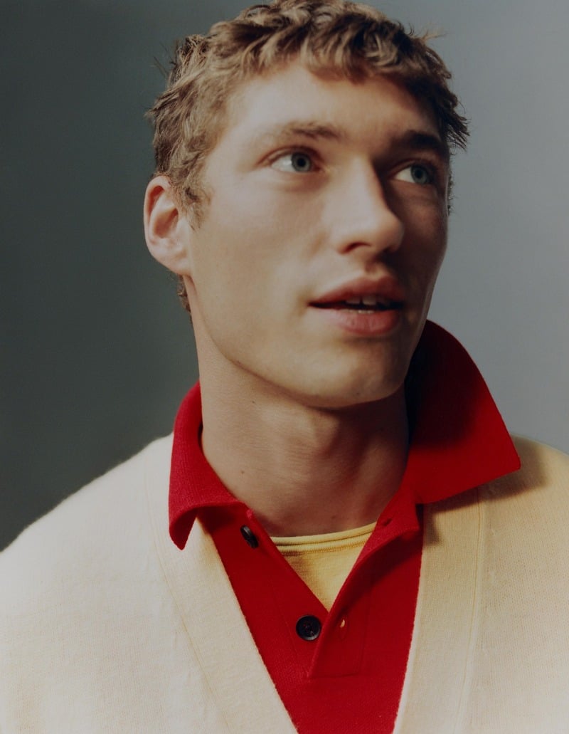 Valentin Humbroich appears in close-up wearing a cream cardigan over a red polo and yellow layer.