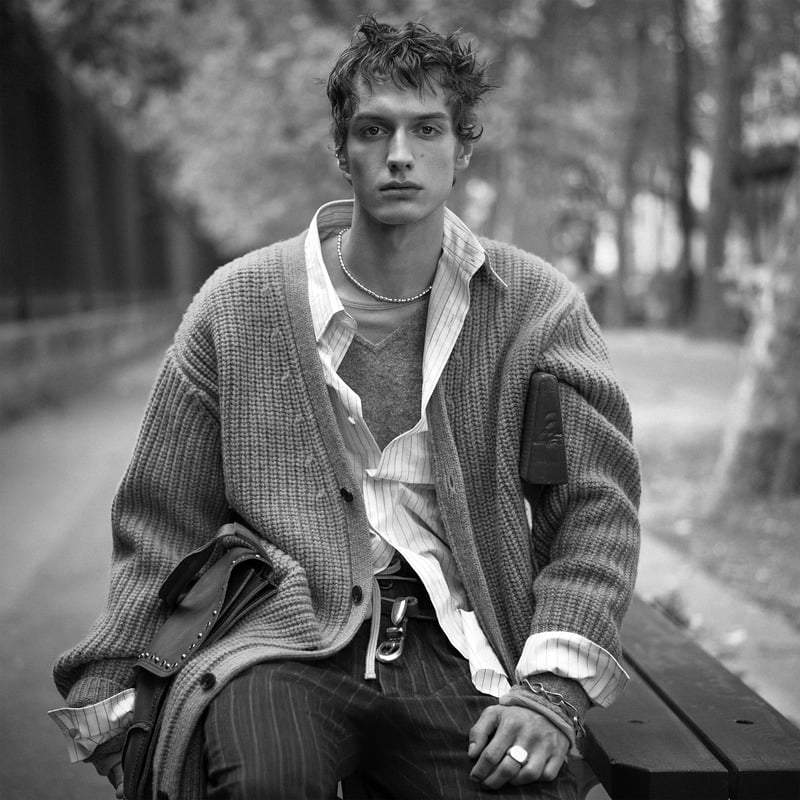 Leonard Kipker sits in a ribbed cardigan over an open striped shirt with a bag on his lap for Zara Studio's fall-winter 2026 campaign
