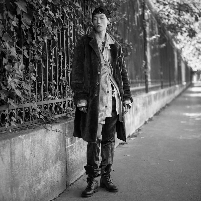 Li Cheng Yuan wears a long overcoat over layered shirting with boots beside an ivy-covered fence for Zara Studio's fall-winter 2026 campaign