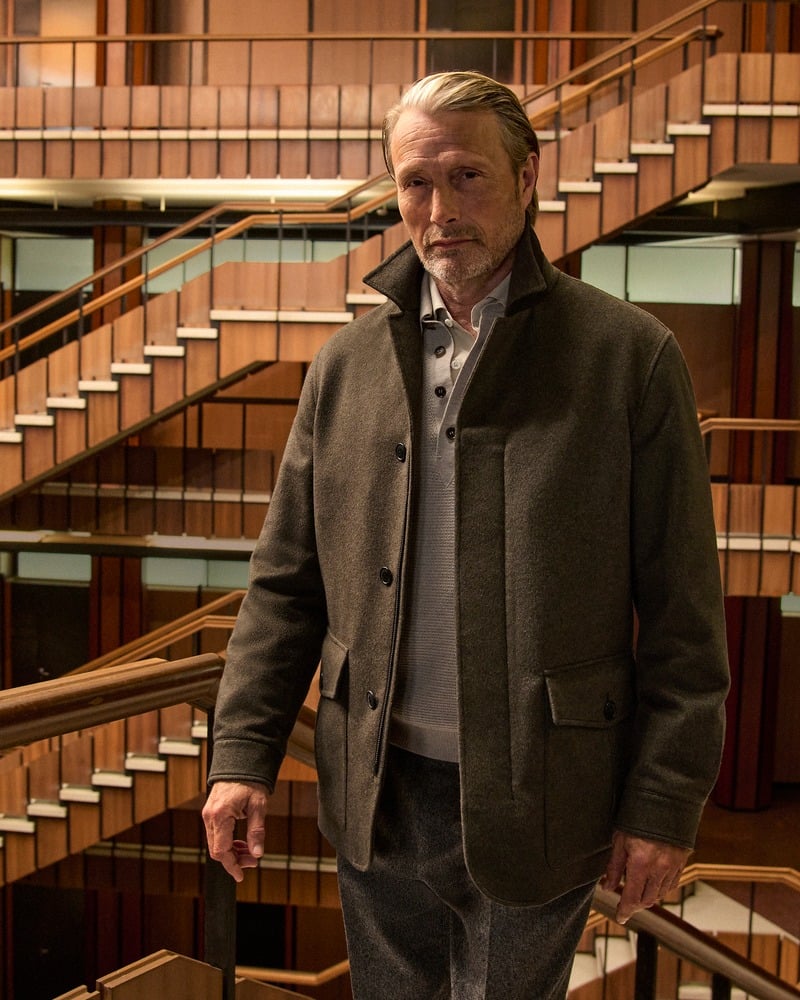 Mads Mikkelsen wears a dark olive Zegna Falcon jacket in a wood-paneled atrium.