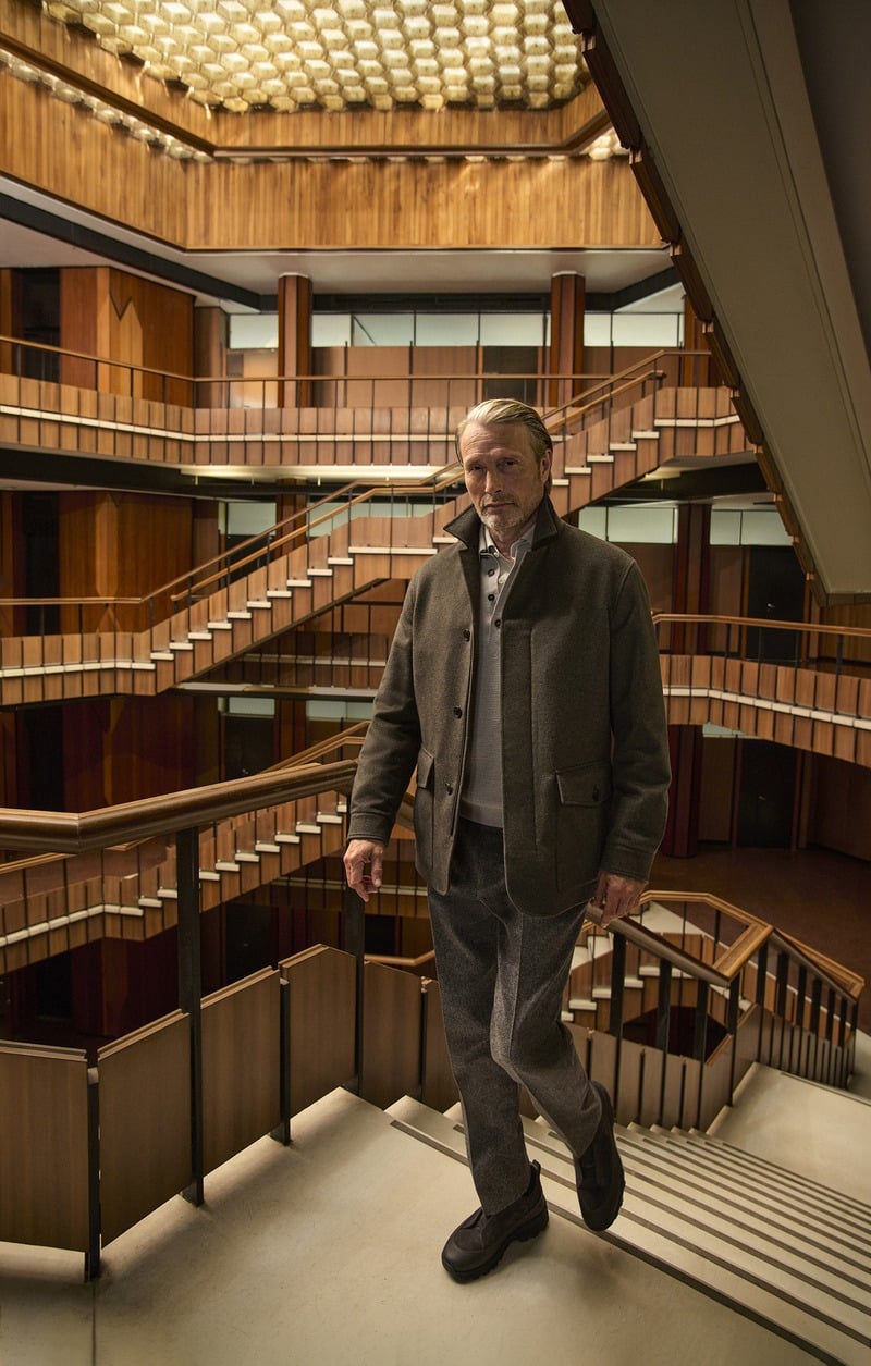 Mads Mikkelsen stands beside an atrium staircase in a dark olive Zegna Falcon jacket and tonal trousers.