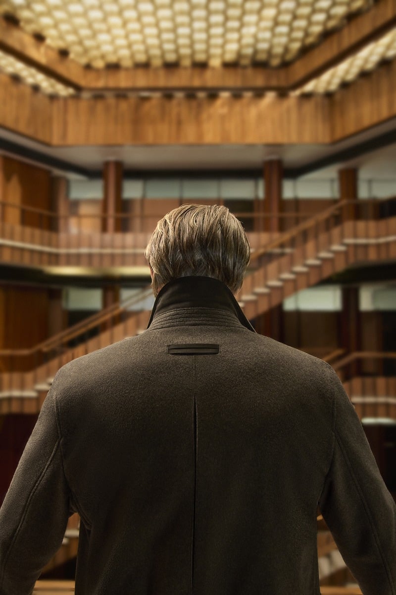 Mads Mikkelsen seen from behind in a dark Zegna jacket in a wood-paneled atrium.