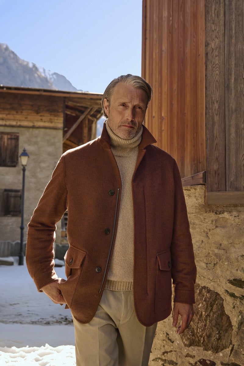 Mads Mikkelsen wears a brown Zegna Falcon jacket over a light turtleneck beside a wooden alpine building.