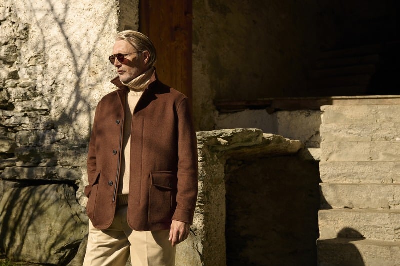 Mads Mikkelsen wears sunglasses and a brown Zegna Falcon jacket beside a stone wall.