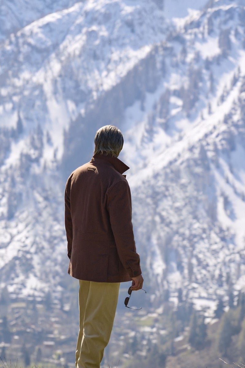 Mads Mikkelsen seen from behind in a brown Zegna Falcon jacket against snowy mountains.