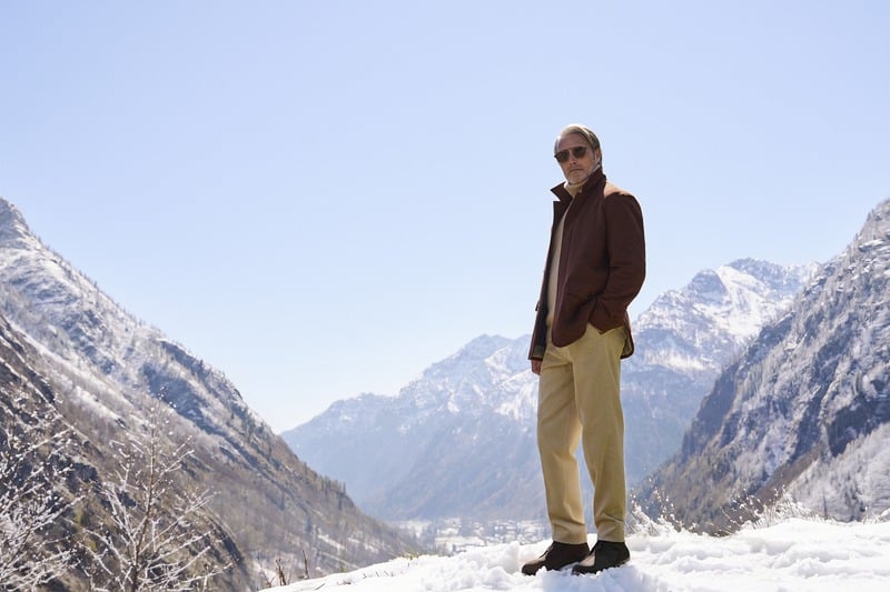 Mads Mikkelsen stands in the snow wearing a brown Zegna Falcon jacket and light trousers.