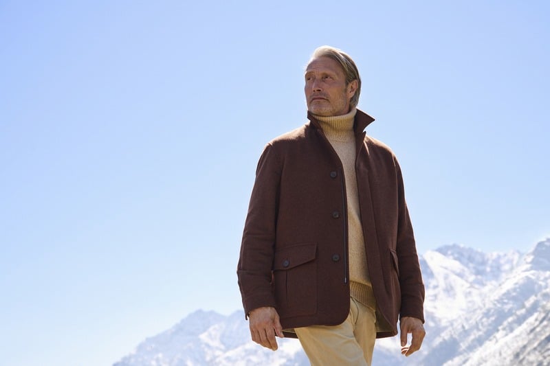 Mads Mikkelsen wears a brown Zegna Falcon jacket over knitwear against a blue alpine sky.