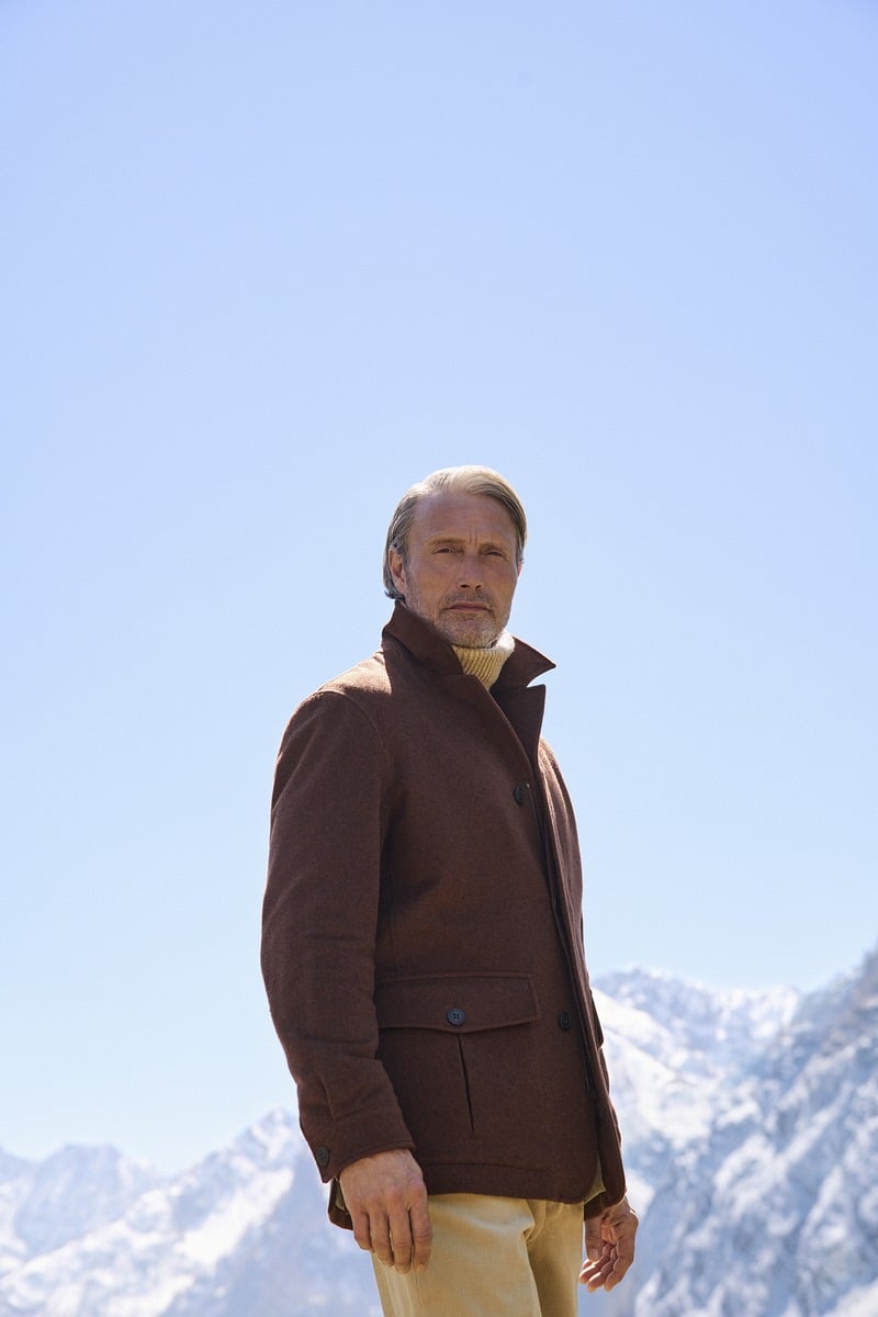 Mads Mikkelsen wears a brown Zegna Falcon jacket and light trousers with snowy peaks behind him.