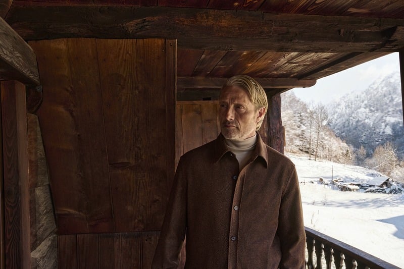 Mads Mikkelsen wears a brown Zegna Falcon jacket on a wooden balcony overlooking snowy mountains.