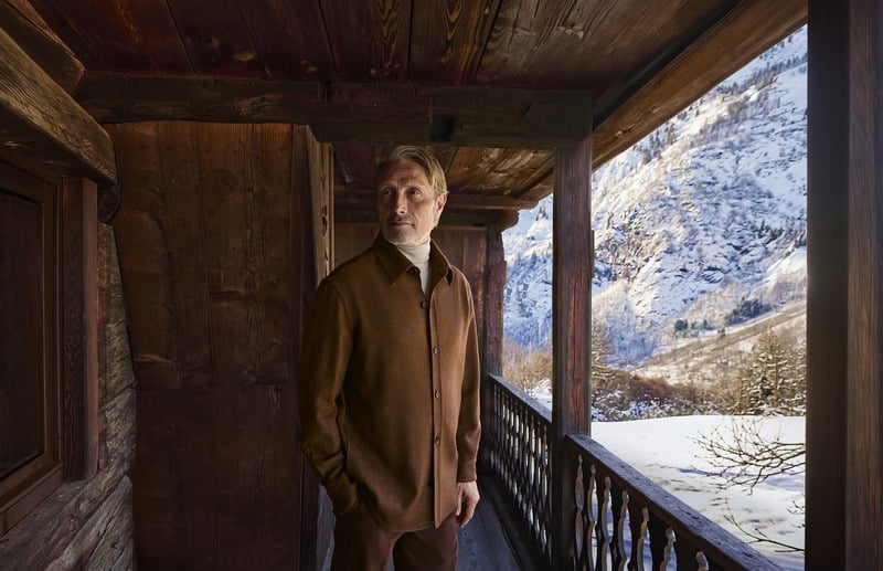 Mads Mikkelsen stands on a wooden balcony in a brown Zegna Falcon jacket over knitwear.
