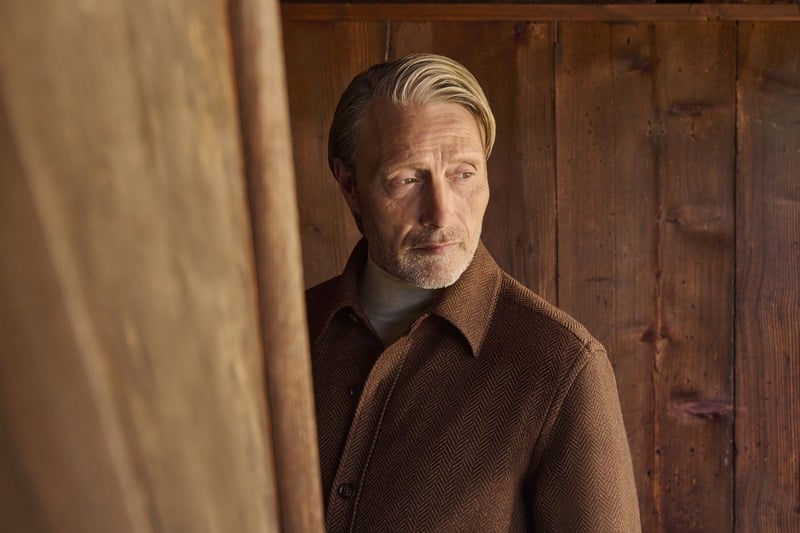 Mads Mikkelsen in a close portrait wearing a brown Zegna Falcon jacket against a wooden wall.